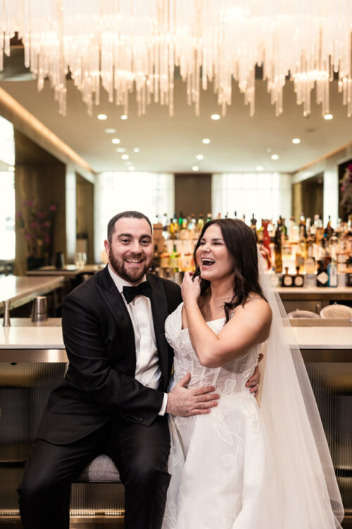 Bride and groom laugh together at a bar in The Four Seasons Chicago, dressed in wedding attire under a modern chandelier