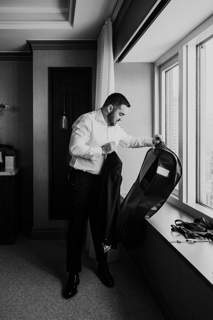 Groom stands by a window at The Four Seasons Chicago, taking a suit jacket from a garment bag while getting ready for his wedding
