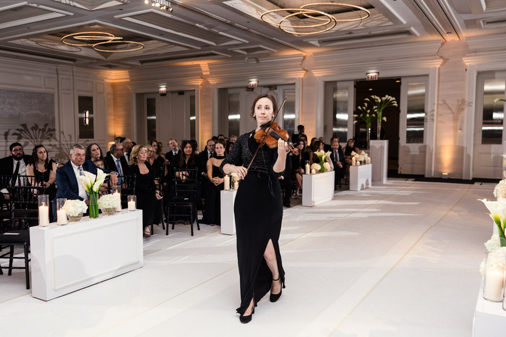 A woman in a black dress plays violin while walking down an aisle during wedding ceremony at The Four Seasons Chicago