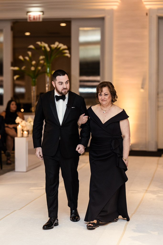 Groom in a tuxedo walks arm-in-arm with his mother in a black dress down the aisle during wedding ceremony at The Four Seasons Chicago
