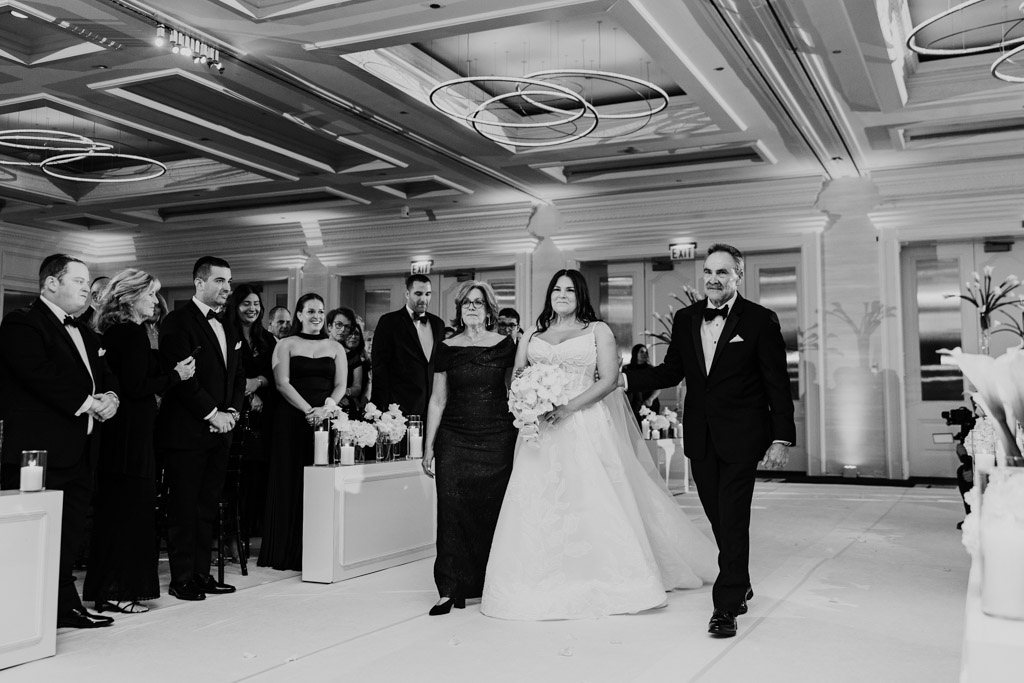 Black and white photo of bride walking down the aisle with her parents as guests watch at The Four Seasons Chicago