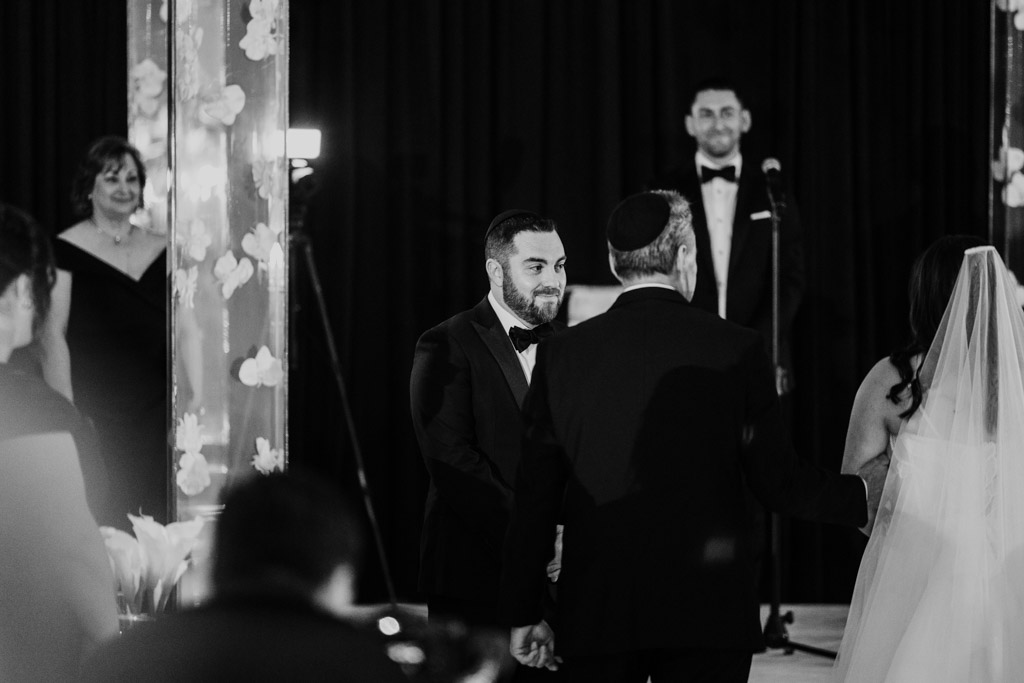 A groom smiles as his bride, in a veil, is walked down the aisle during their Four Seasons Chicago wedding ceremony