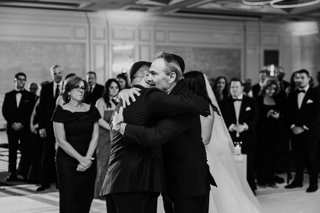 A groom and an older man hug at a formal wedding ceremony at The Four Seasons Chicago as guests look on, all dressed in black-tie attire.