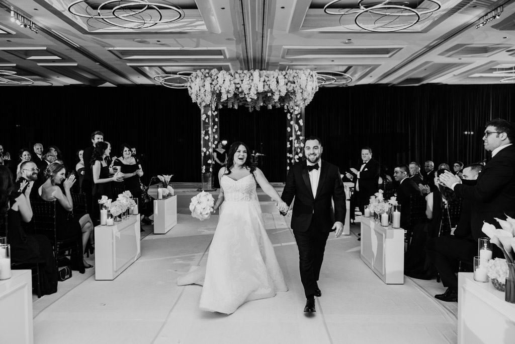 Black and white photo of bride and groom walking down the aisle, smiling, as guests cheer and applaud during their wedding ceremony at The Four Seasons Chicago