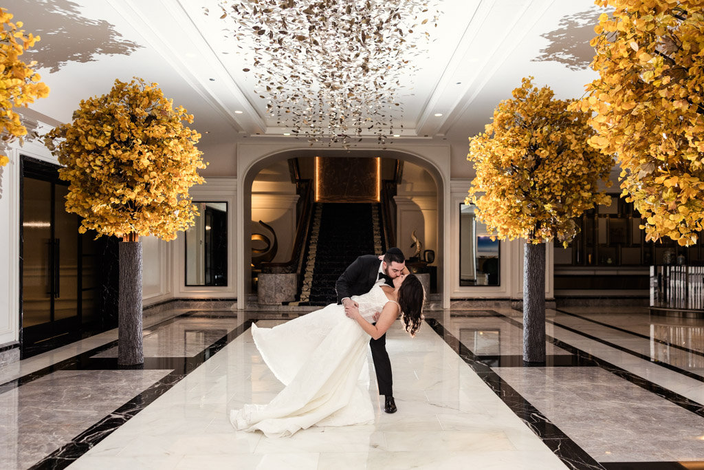 A groom dips his bride for a kiss in an elegant hall with golden trees and marble floors at The Four Seasons Chicago.