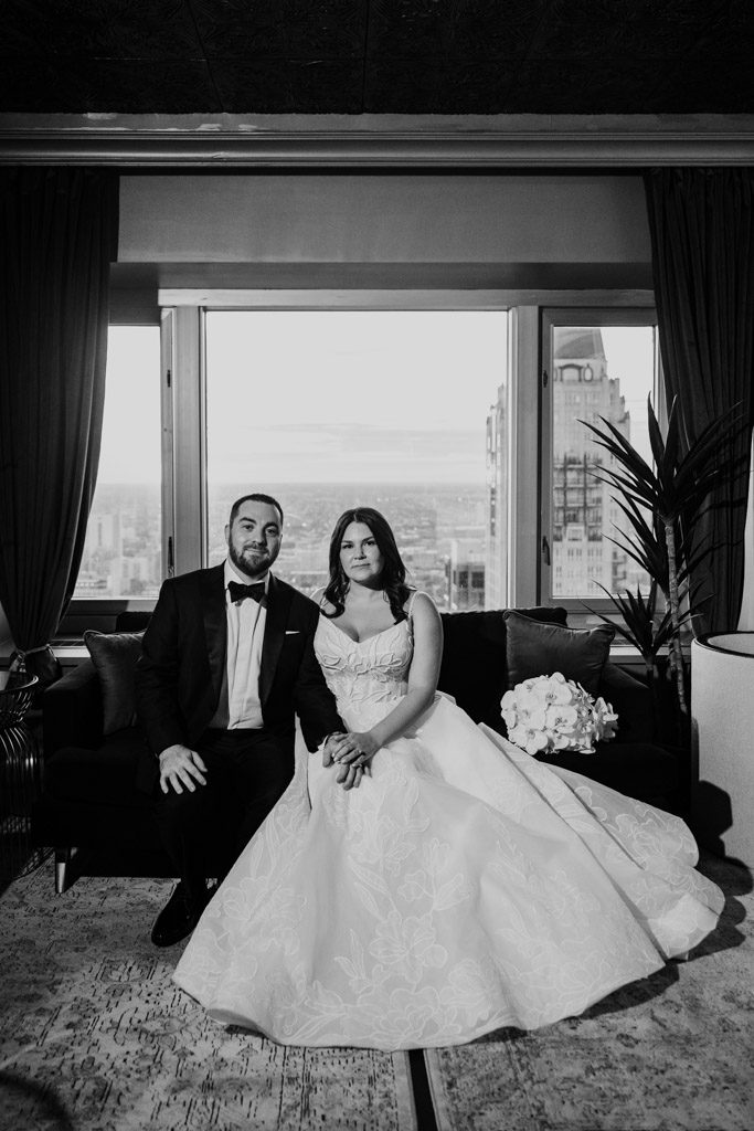 A bride and groom sit on a couch in front of a large window with a city view at The Four Seasons Chicago, dressed in formal attire.