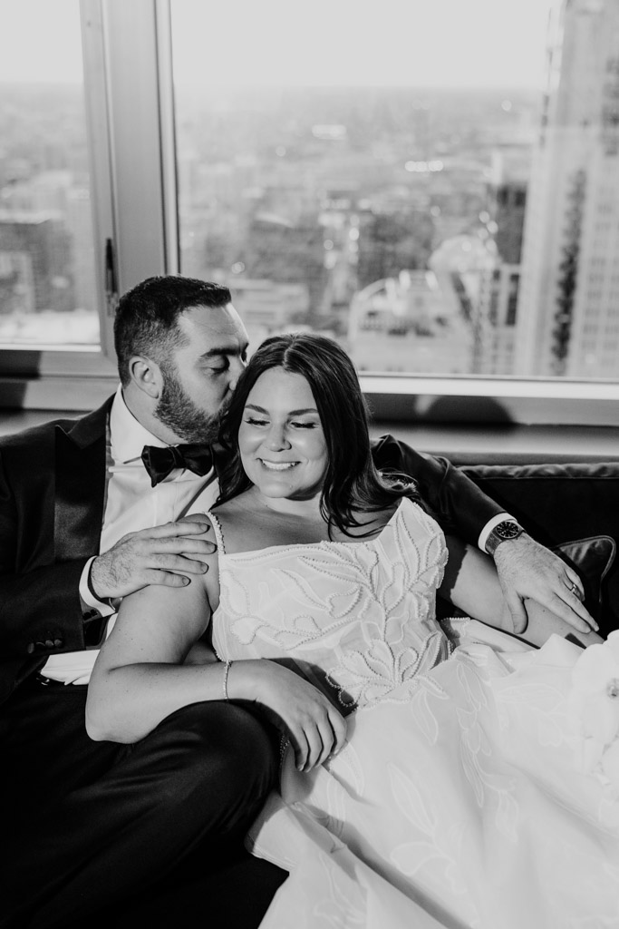 Black and white photo of groom kissing his bride on the head as they sit together, smiling, in front of a cityscape window at The Four Seasons Chicago