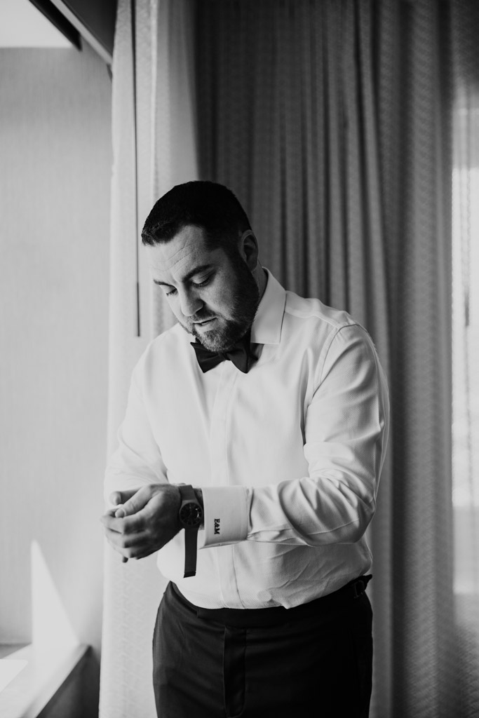 Black and white photo of groom adjusting his cufflink while wearing a white shirt and bow tie, standing by a window at The Four Seasons Chicago