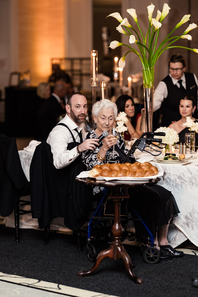 Bride's grandmother with a walker speaks into a mic before slicing the ceremonial challah bread during weding reception at The Four Seasons Chicago