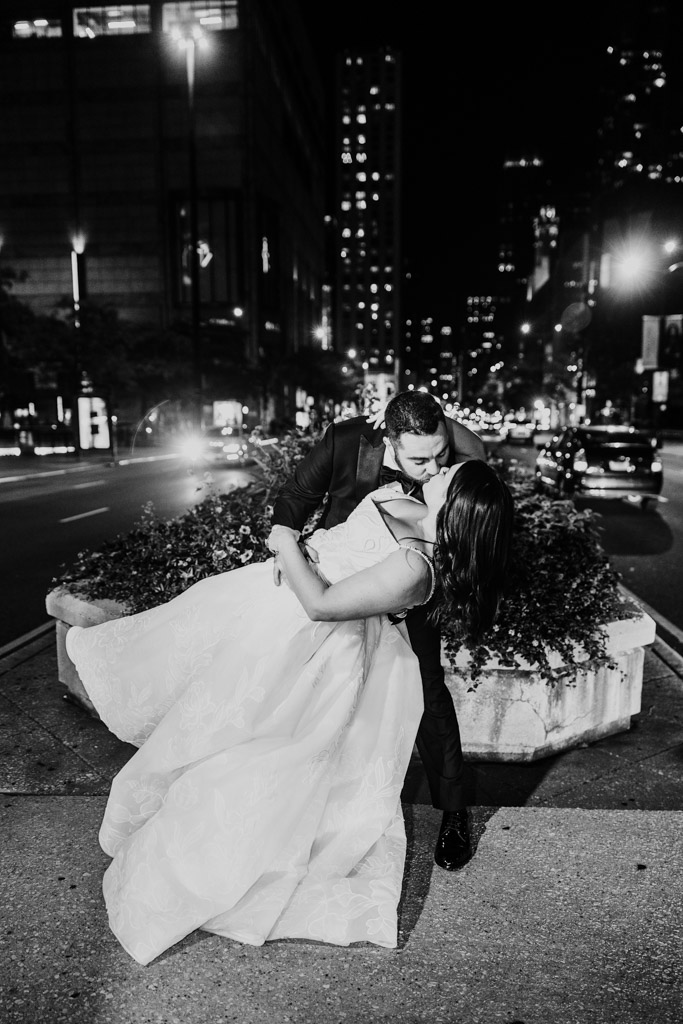 Black and white photo of newlywed couple in formal attire kissing on a downtown Chicago street at night