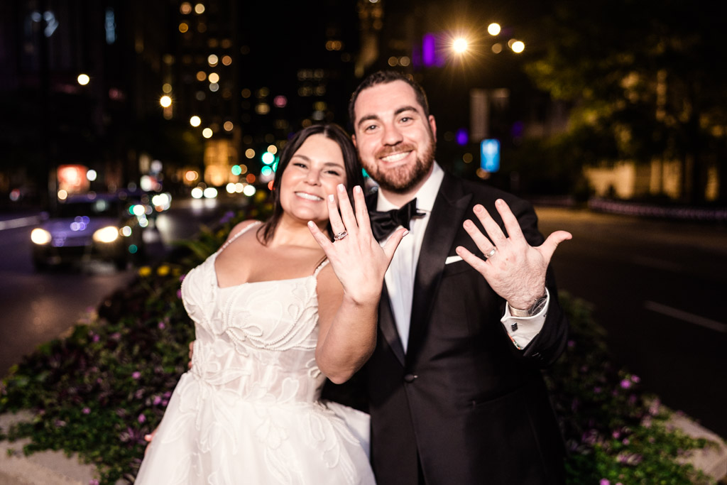 Bride and groom smiling at night on a city street, holding up hands to show their wedding rings after celebrating at The Four Seasons Chicago