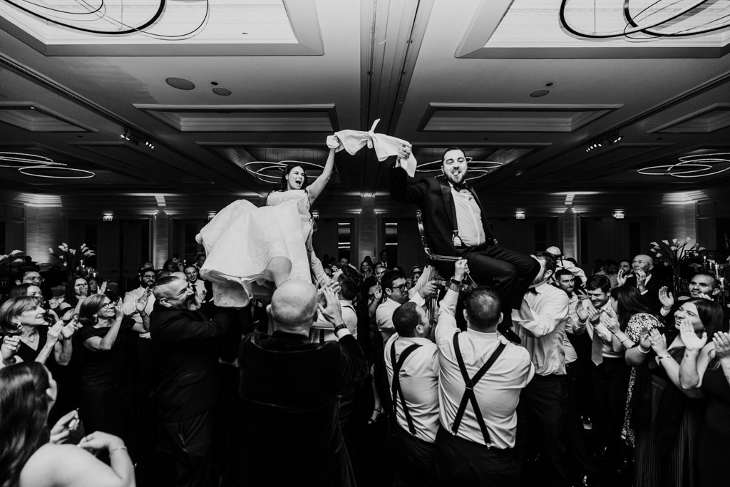 A bride and groom are lifted on chairs by guests during the hora as part of their wedding reception at The Four Seasons Chicago