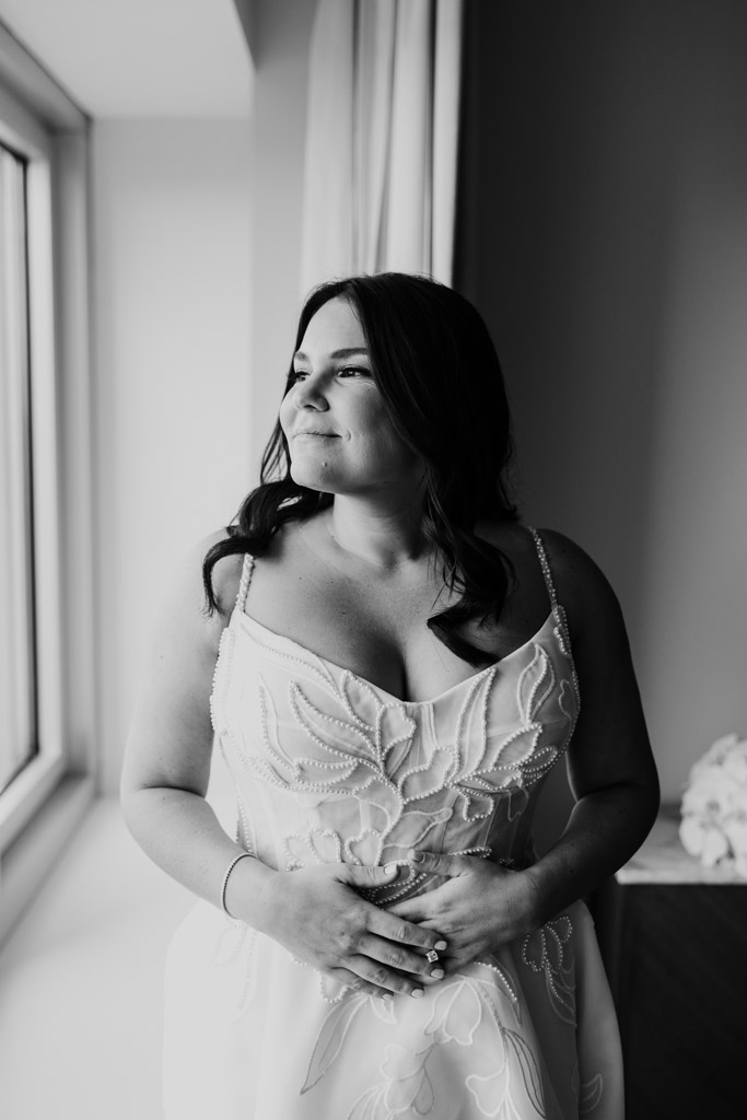 Black and white photo of woman in a wedding dress standing by a window at The Four Seasons Chicago, looking outside with a gentle smile