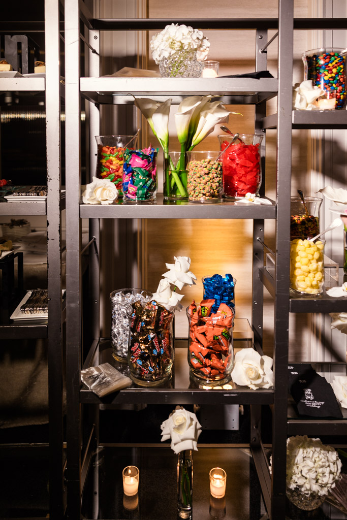 A metal shelf at The Four Seasons Chicago wedding reception holds jars of colorful candy, white flowers, and two lit candles at the bottom