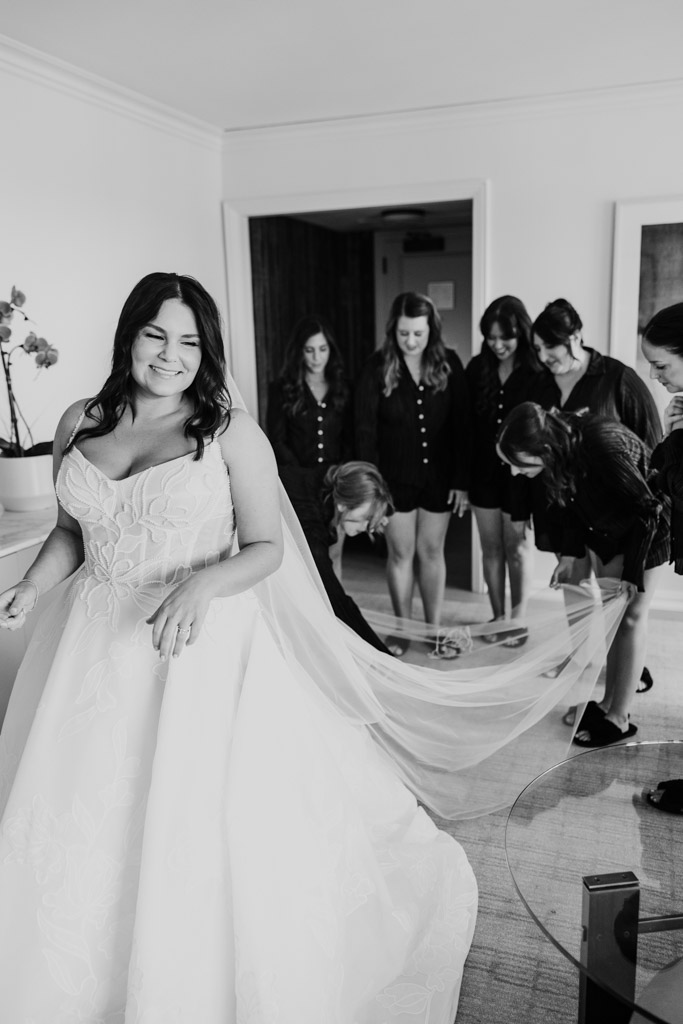 Black and white phot of bride in a wedding dress smiling as bridesmaids adjust her train in a bright room at The Four Seasons Chicago
