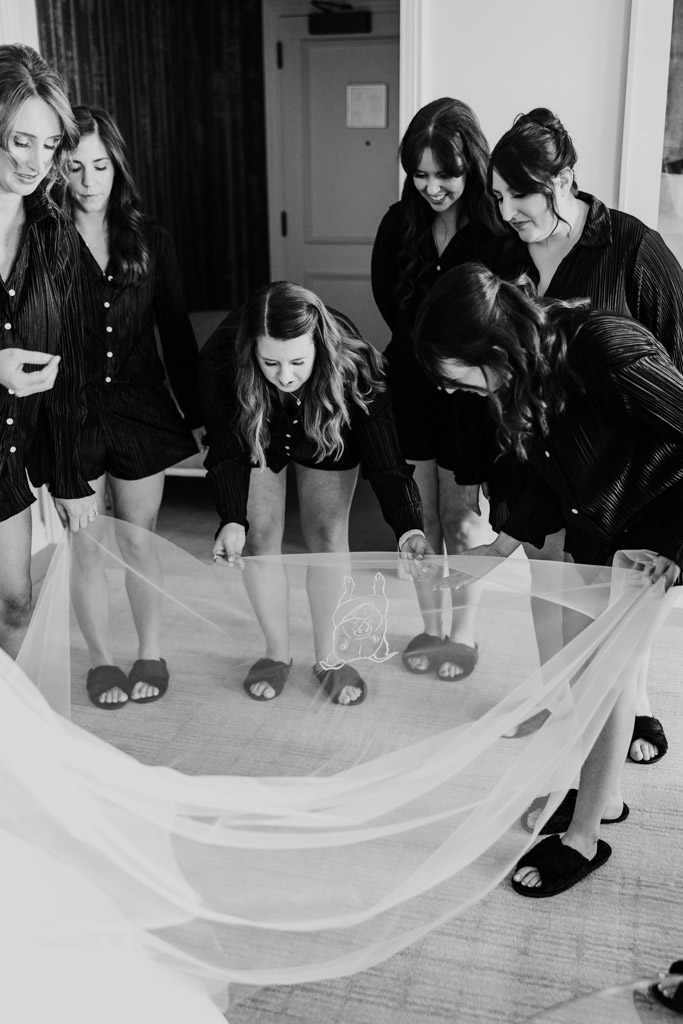 Six bridesmaids in matching outfits smile while holding and admiring the bride's custom embroidered veil in a bright room at The Four Seasons Chicago