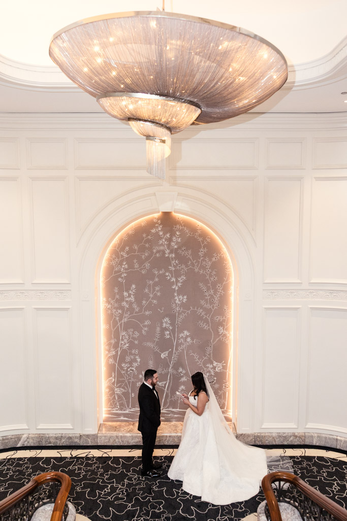 A bride and groom face each other beneath a large chandelier in an elegant, white-walled room at The Four Seasons Chicago