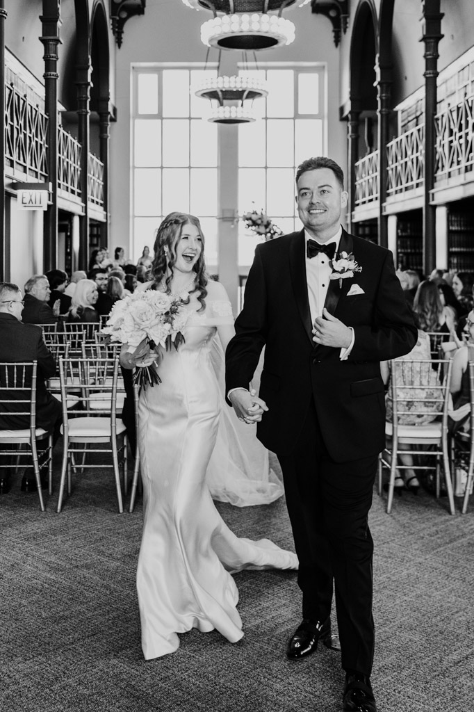 A bride and groom walk down the aisle smiling, holding hands, in a formal indoor wedding ceremony.