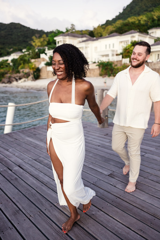Happy newlywed couple walks barefoot on a wooden pier by the beach at Viceroy Sugar Beach, holding hands and wearing white outfits