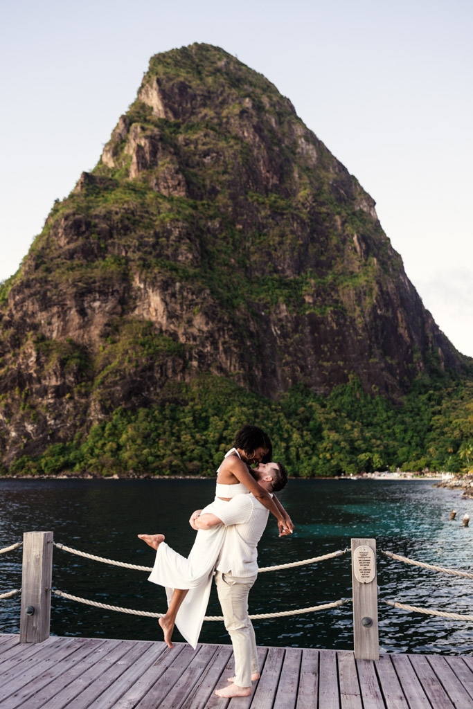 Man lifts his partner as they stand on a dock at Viceroy Sugar Beach in St. Lucia embraces on a dock at Viceroy Sugar Beach, with Petit Piton and the ocean in the background.
