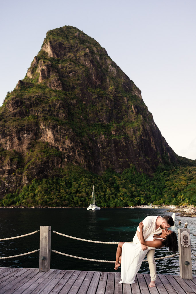 Newlywed couple kisses on a wooden dock at Viceroy Sugar Beach, with a sailboat and a tall, lush mountain in the background