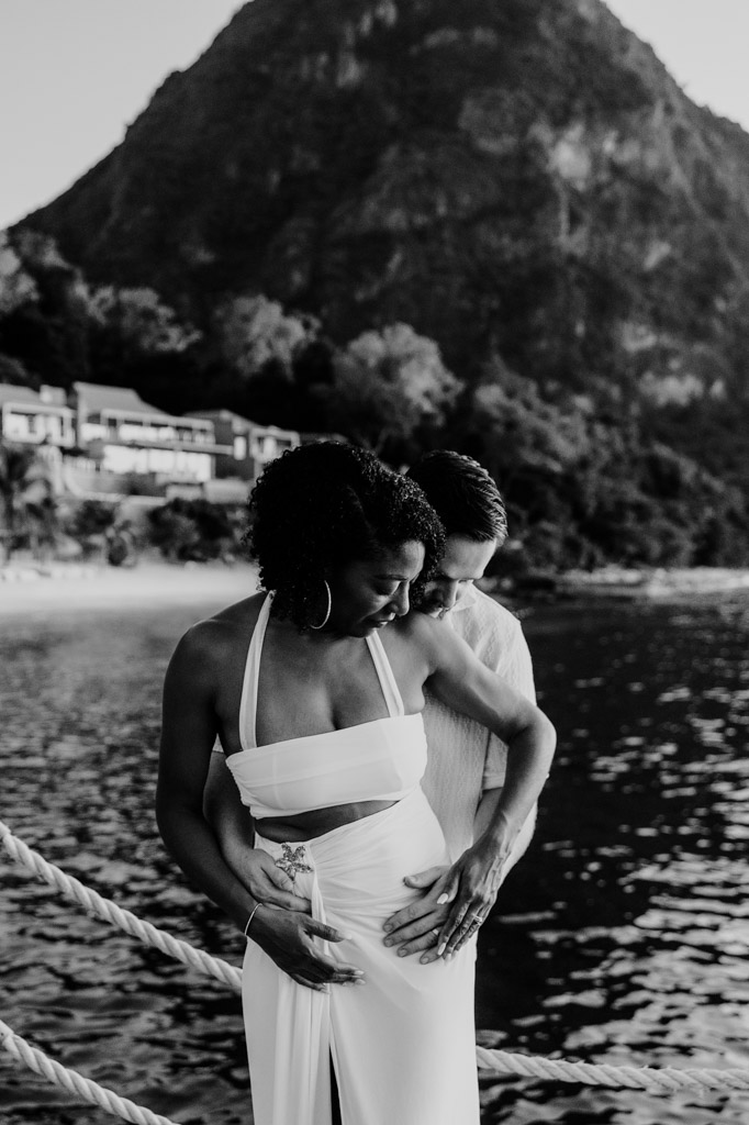 Black and white photo of newlywed couple by the water with Gros Piton and villas in the background at Viceroy Sugar Beach