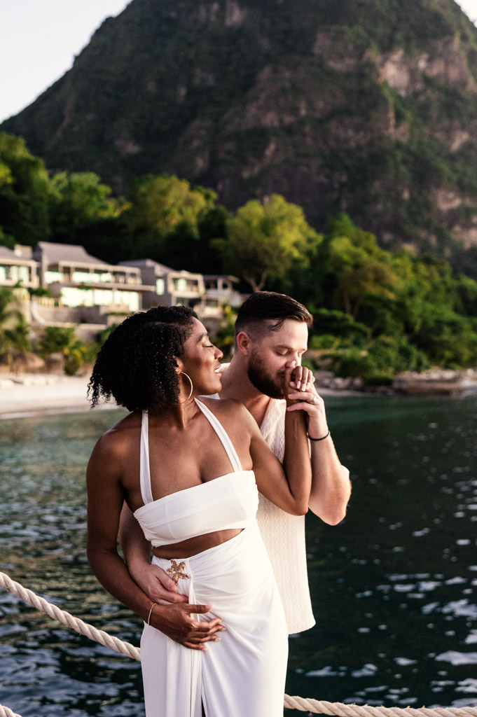 Man kisses his partner's hand as they stand on a pier by the water, with Gros Piton and Viceroy Sugar Beach villas in the background