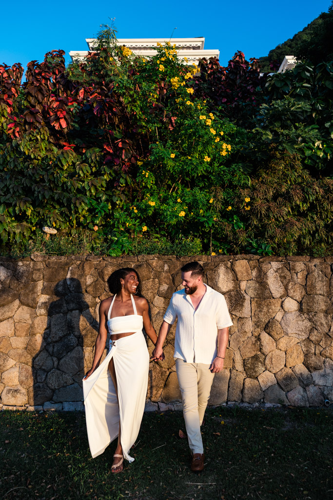 Happy newlywed couple in light outfits holds hands, walking by a stone wall with lush greenery and flowers at Viceroy Sugar Beach