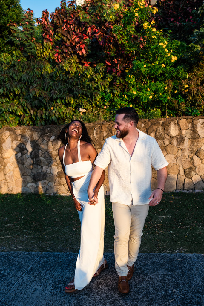 A smiling couple holding hands and walking outdoors by a stone wall with lush greenery in the background at Viceroy Sugar Beach