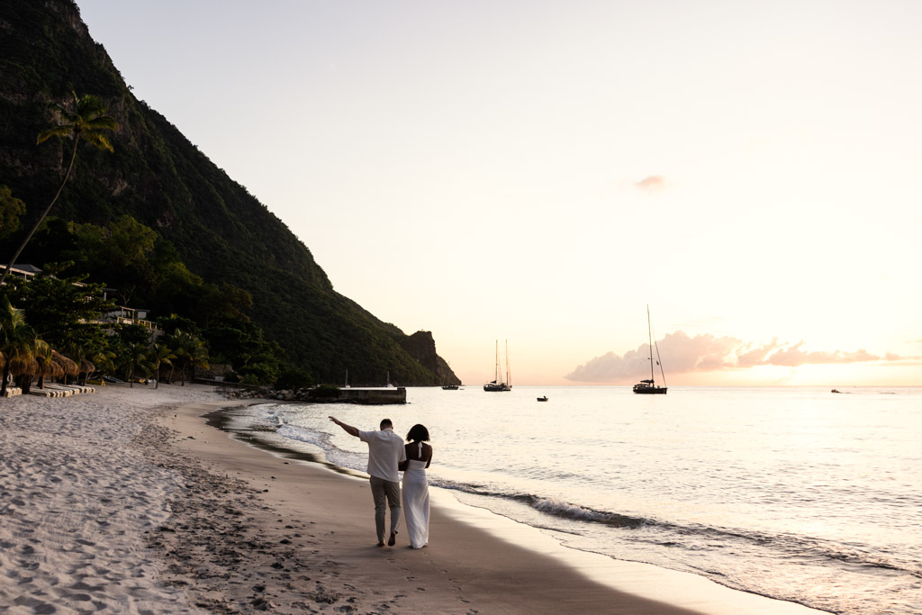 A couple walks on a sandy beach at sunset, with sailboats and Gros Piton mountain behind them at Viceroy Sugar Beach in St. Lucia