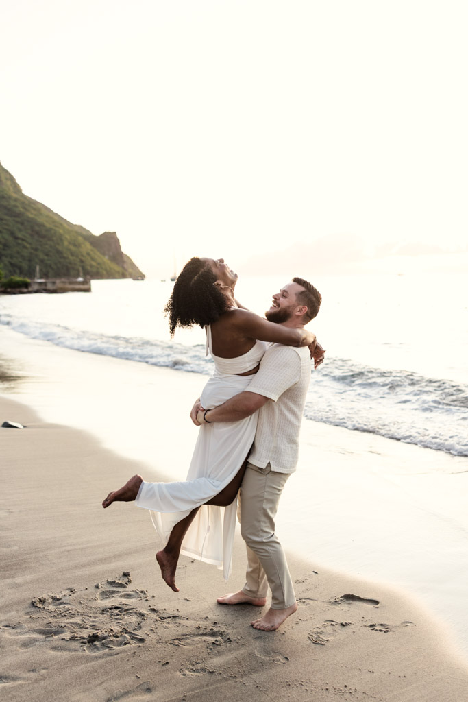 A man lifts his partner joyfully on the sandy shores of Viceroy Sugar Beach at sunset, both smiling and barefoot by the ocean