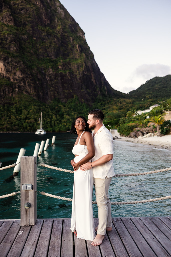 A couple embraces on a wooden dock by turquoise water at Viceroy Sugar Beach, with Petit Piton and a sailboat in the background