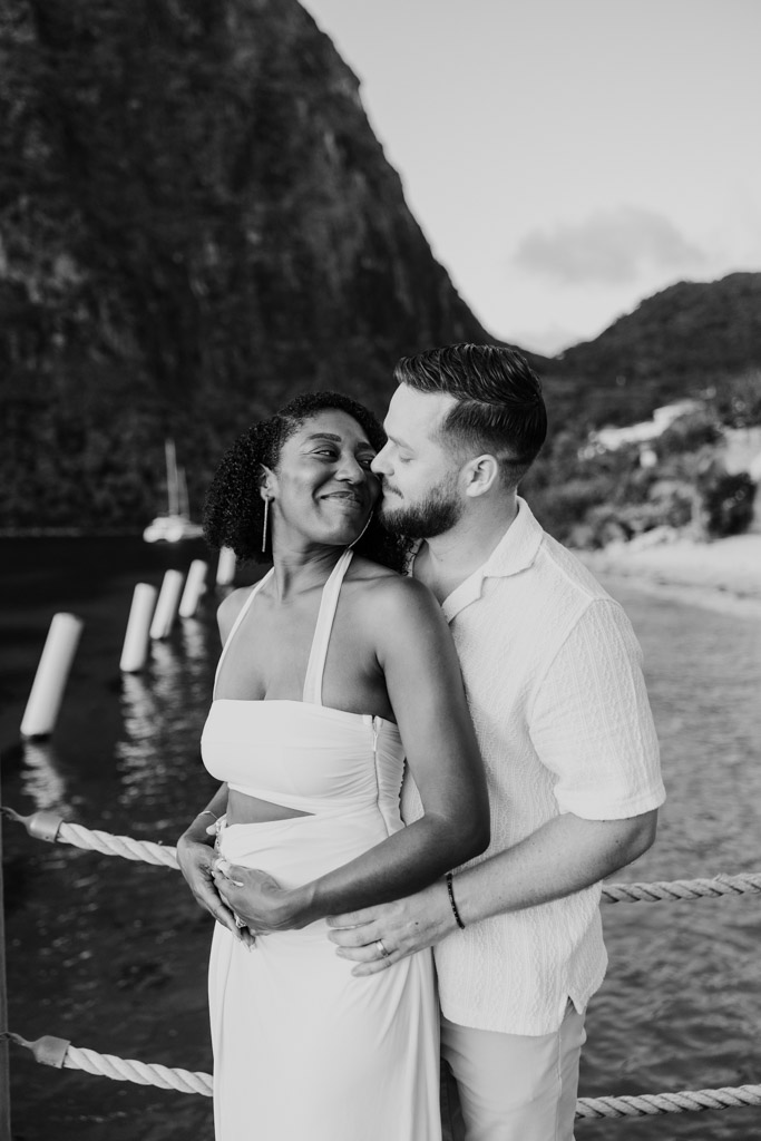 Black and white photo of newlywed couple embracing and smiling at each other by the water, with mountains in the background at Viceroy Sugar Beach