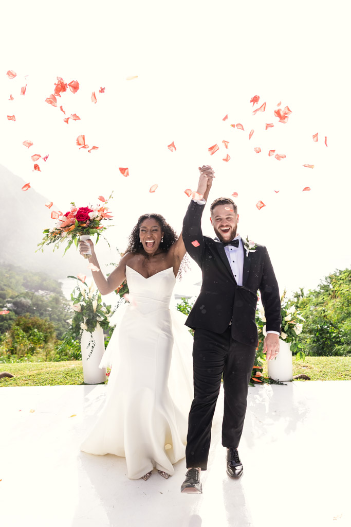 A joyful bride and groom celebrate outdoors under falling rose petals, holding hands and smiling.