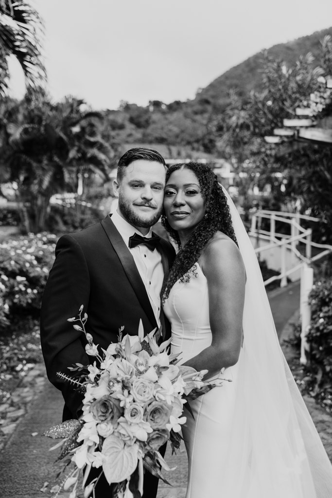 Bride and groom in wedding attire pose outdoors, holding a bouquet, with greenery in the background.