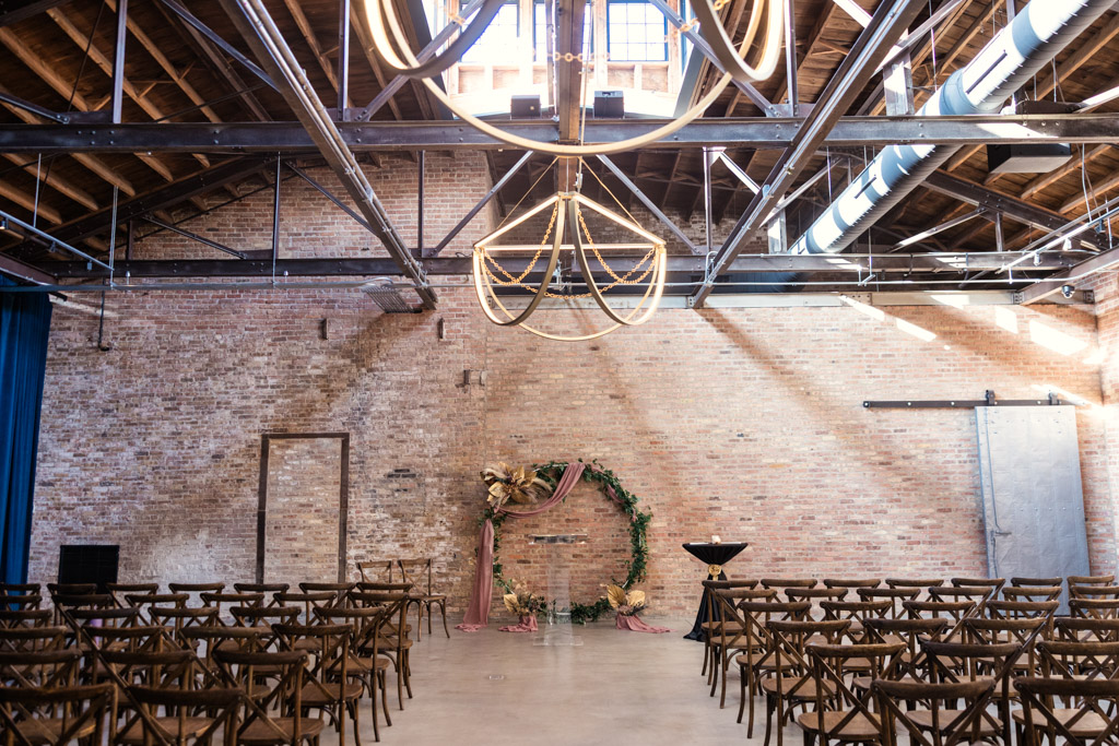 Industrial-style Wildman BT wedding venue with wooden chairs, brick walls, and a circular floral arch beneath modern chandeliers.
