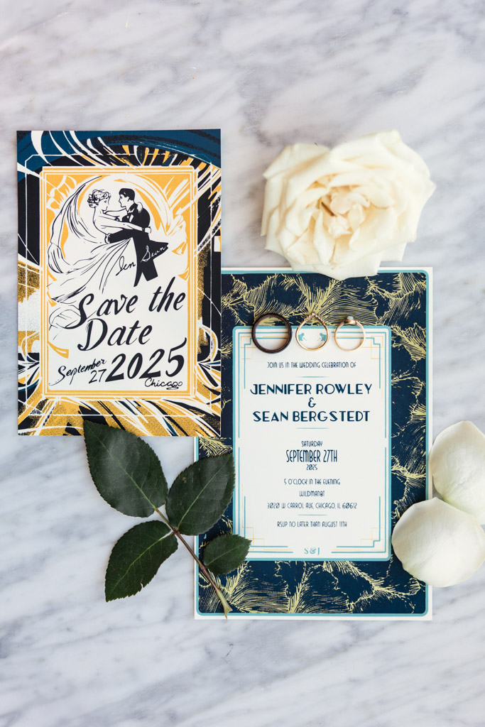 Two wedding invitations, two rings, and white rose petals rest on a marble surface—elegant details perfect for a Wildman BT wedding celebration.