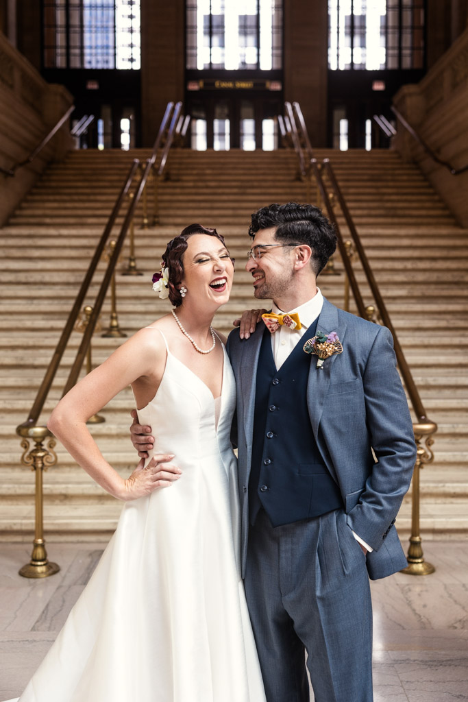 Bride and groom smiling and laughing, standing together in front of a grand staircase indoors at their unforgettable Wildman BT wedding.