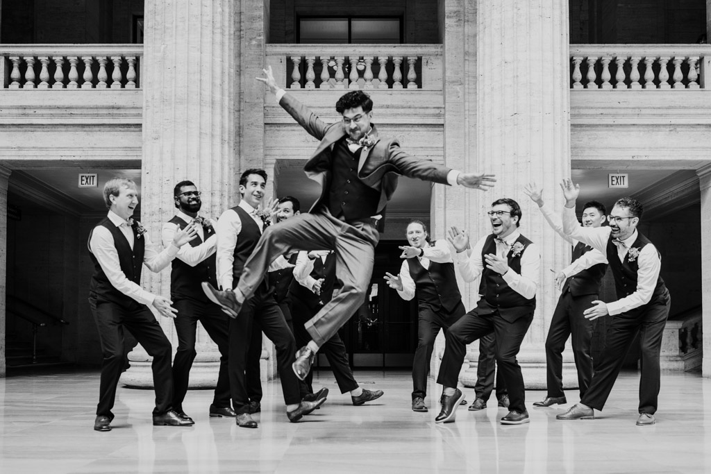 A groom jumps mid-air as his groomsmen cheer and gesture around him in a grand hall during a lively Wildman BT wedding celebration.