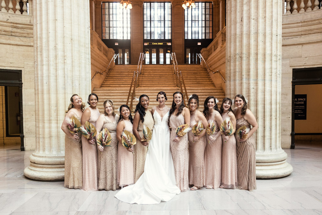 A bride with her bridesmaids in blush dresses pose in front of grand marble columns and stairs indoors at a stunning Wildman BT wedding.
