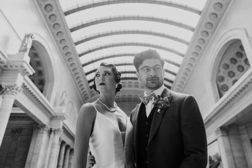 Moody black and white portrait of bride and groom standing together in Union Station with arched skylights above them