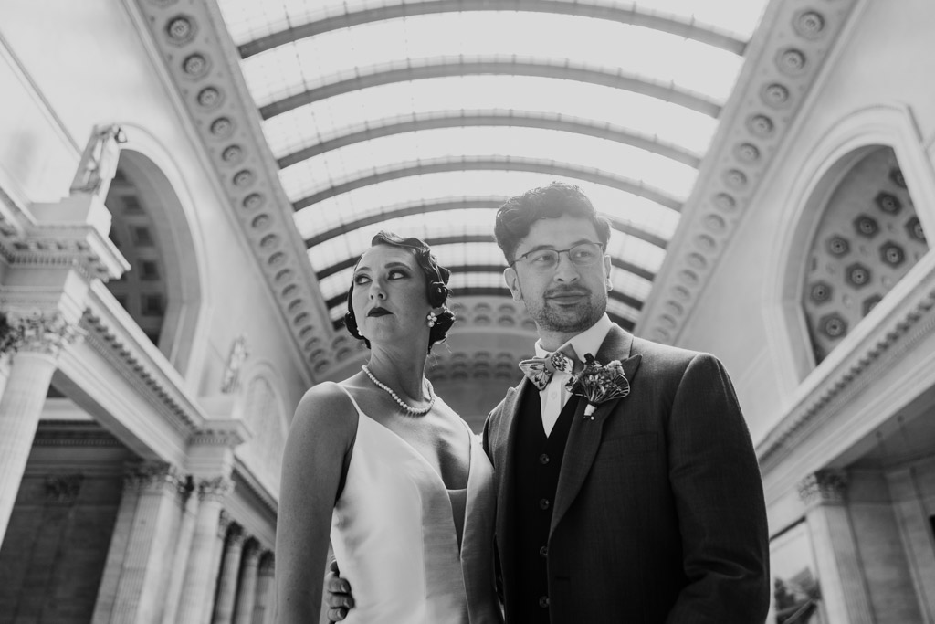 A bride and groom stand together in a grand, ornate hall with arched windows above, capturing the elegance of their Wildman BT wedding.