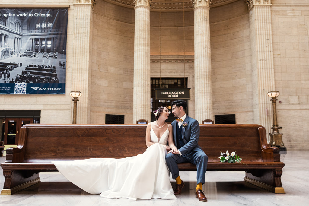A Wildman BT wedding bride and groom sit close together on a wooden bench in a grand, elegant hall with tall columns.