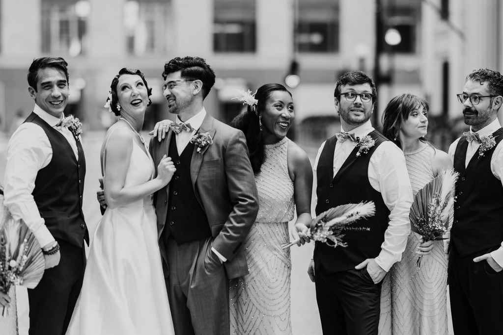 Six people in wedding attire smile and laugh together outdoors in a city, holding bouquets—capturing joyful moments from the Wildman BT wedding.