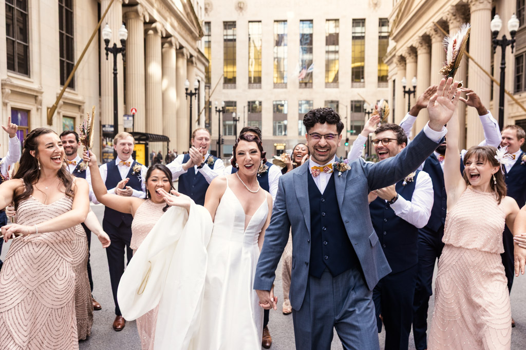 A joyful Wildman BT wedding party celebrates in formal attire on a city street, with the bride and groom in front.