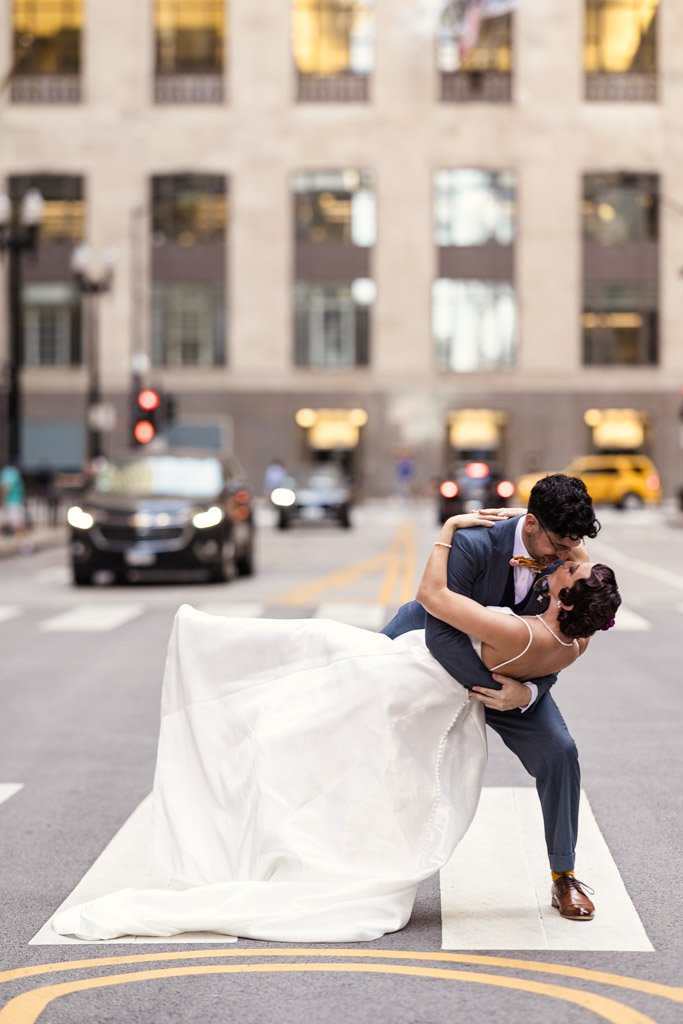 A groom dips and kisses his bride in the middle of a city street, surrounded by traffic and tall buildings, capturing a bold Wildman BT wedding moment.