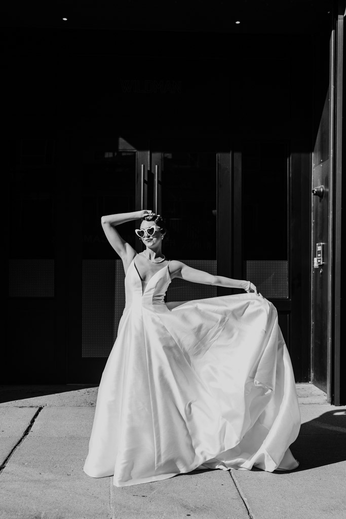A woman in a flowing Wildman BT wedding dress and sunglasses poses confidently on a sidewalk in front of a building.