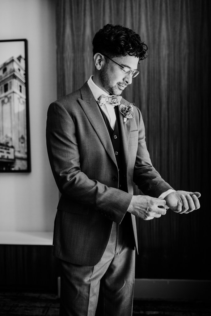 A man in a suit adjusts his cufflinks, standing indoors and smiling, in a black and white photo—capturing a timeless moment before the Wildman BT wedding.