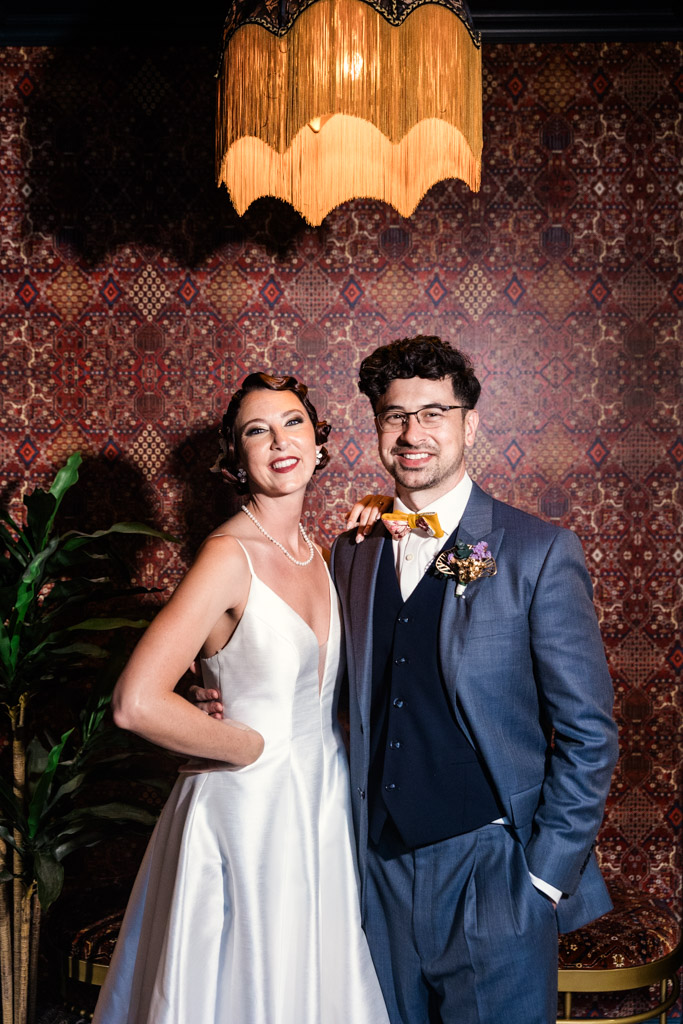 A Wildman BT wedding: A bride and groom in formal attire smile together under a vintage lamp with a patterned wallpaper background.