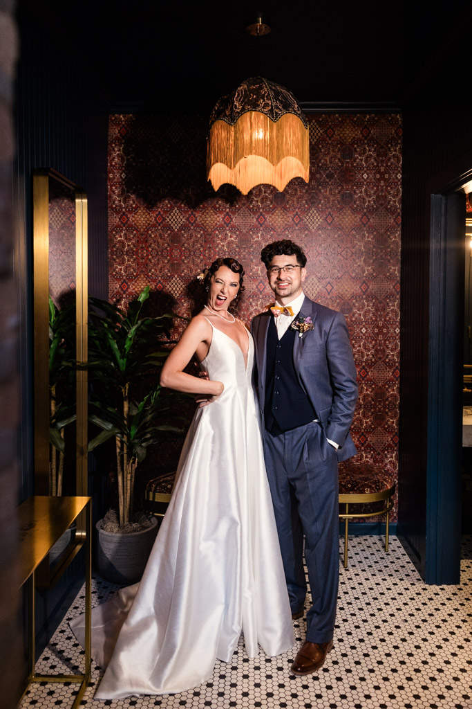 Bride in a white dress and groom in a blue suit pose and smile together at their Wildman BT wedding in a stylish, vintage-themed room.
